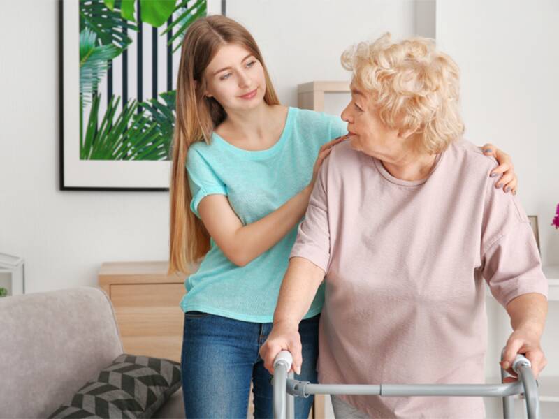 Eine junge Frau unterstützt eine ältere Frau mit einem Rollator in einem freundlich eingerichteten Wohnzimmer.