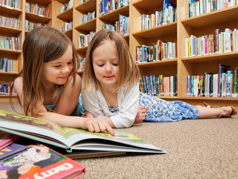 Zwei junge Mädchen liegen auf dem Boden und lesen gemeinsam ein Buch in einer Bibliothek mit Bücherregalen im Hintergrund.