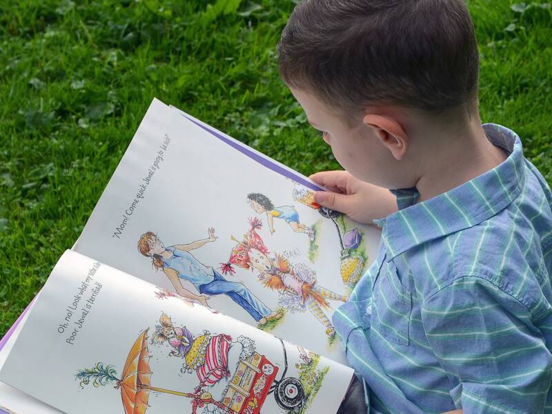 Ein Kind sitzt auf grünem Gras und liest ein aufgeschlagenes Bilderbuch, während es ein rot-weißes Schuhwerk trägt.