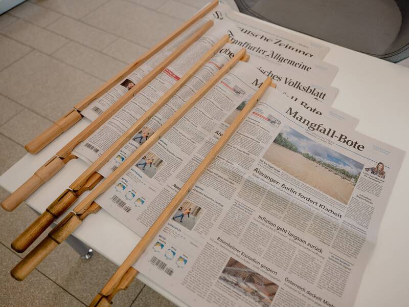 Vier aufgeschlagene Zeitungen liegen auf einer weißen Oberfläche, gehalten von vier hölzernen Zeitungsständern.