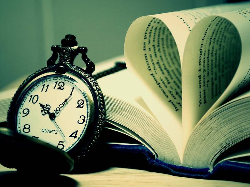 Ein offenes Buch mit gespiegelten Seiten, die ein Herz formen, liegt neben einer alten Taschenuhr auf einem Tisch.