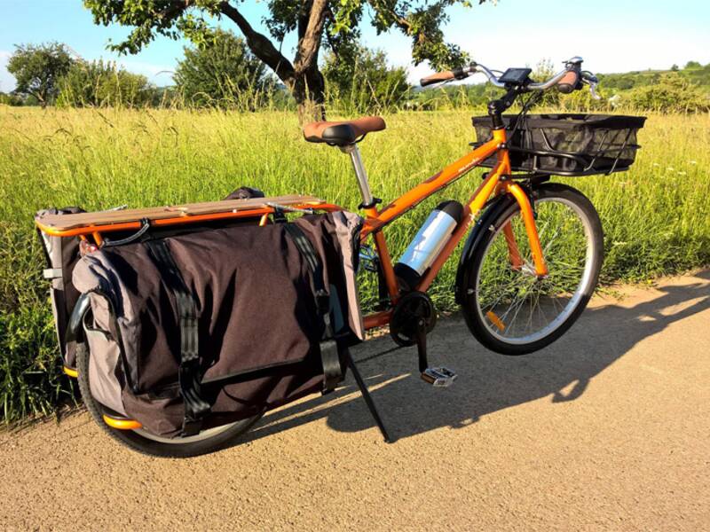 Ein orangefarbenes Lastenfahrrad mit großen Seitentaschen steht auf einem Weg am Rand eines Feldes bei sonnigem Wetter.