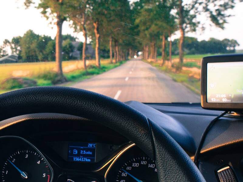 Fahrersitz und Lenkrad im Innenraum eines Autos mit Blick auf eine Straße mit Bäumen.