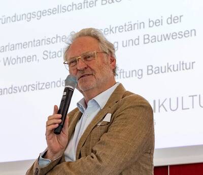 Dr. Ernst Böhm hält ein Mikrofon und spricht vor einer großen Leinwand mit Text.
