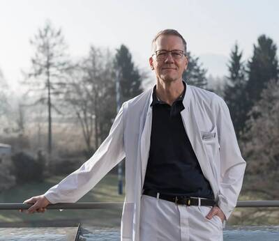 Dr. Alex Höfter steht in einem weißen Kittel draußen vor einer Landschaft mit Bäumen.