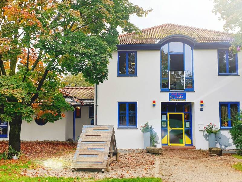 Das Bild zeigt ein zweistöckiges Gebäude mit einem kreisförmigen Dach und bunten Fensterrahmen, umgeben von Bäumen mit herbstlich gefärbtem Laub.
