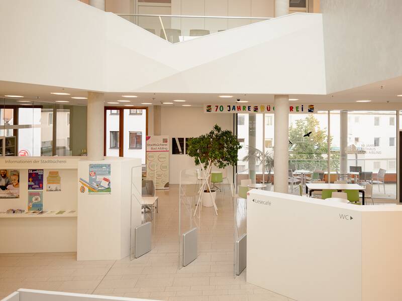 Ein moderner, heller Bibliotheksraum mit Informationsbereich, Pflanzen und Sitzgelegenheiten, dekoriert mit einem Banner "70 Jahre Bücherei".