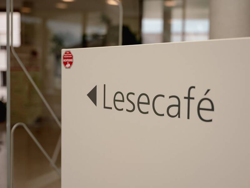 Ein Schild mit der Aufschrift "Lesecafé" zeigt nach links und ist im Inneren eines modernen Gebäudes angebracht.