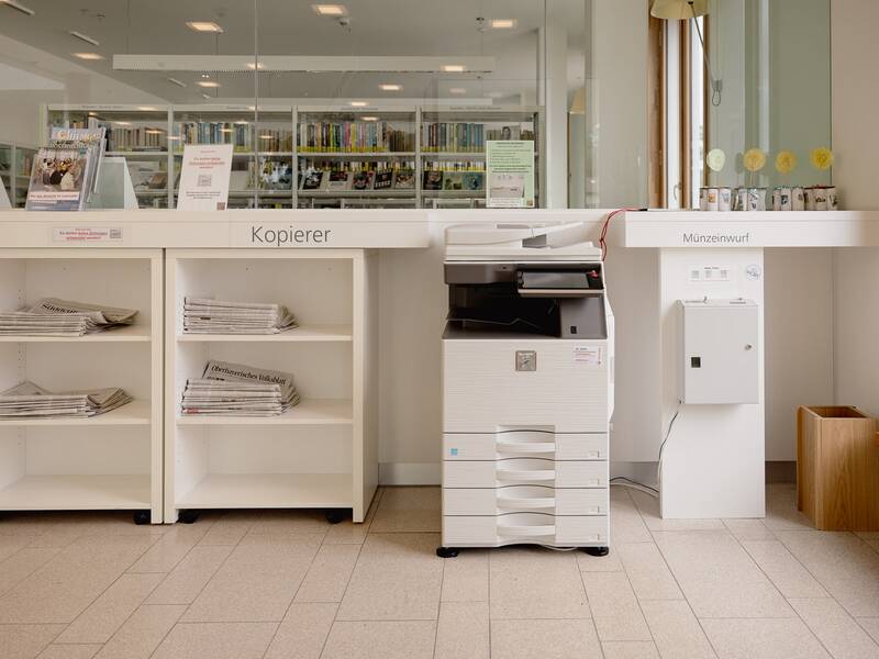 Ein weißer Kopierer steht in einer Bibliothek neben Regalen mit Zeitungen und Münzeinwurf.