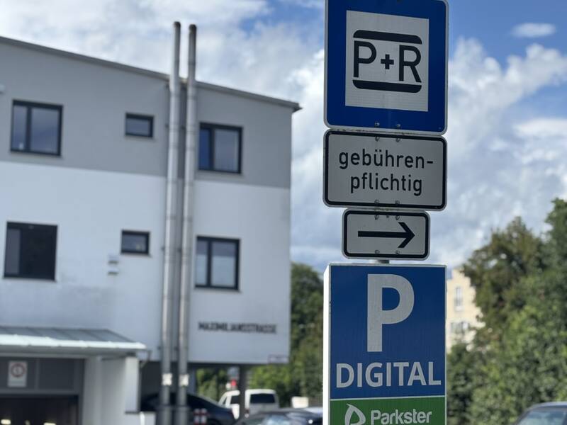 Ein Parkplatzschild mit der Aufschrift "P+R gebührenpflichtig" und "P Digital" steht vor einem weißen Gebäude bei sonnigem Wetter.