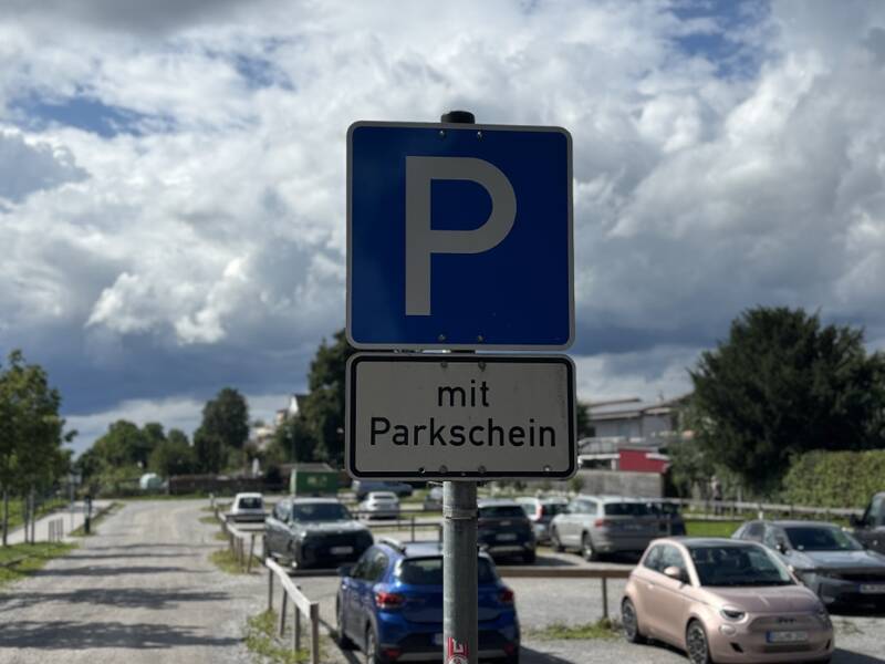 Ein Parkplatzschild mit der Aufschrift "mit Parkschein" steht vor einem Parkplatz voller Autos unter einem bewölkten Himmel.