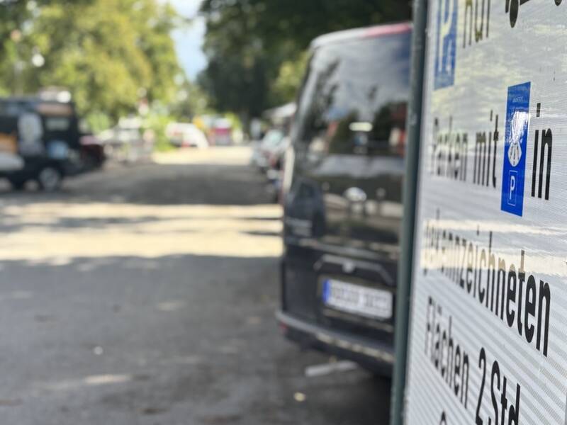 Ein vergrößertes Parkplatzschild im Vordergrund zeigt Parkbedingungen für Fahrzeuge, während im Hintergrund Autos auf einer von Bäumen gesäumten Straße geparkt sind.