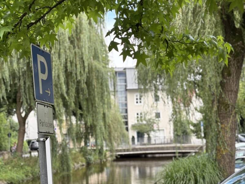 Ein Parkplatzschild befindet sich neben einem ruhigen Fluss, umgeben von üppigem Grün und geparkten Autos.
