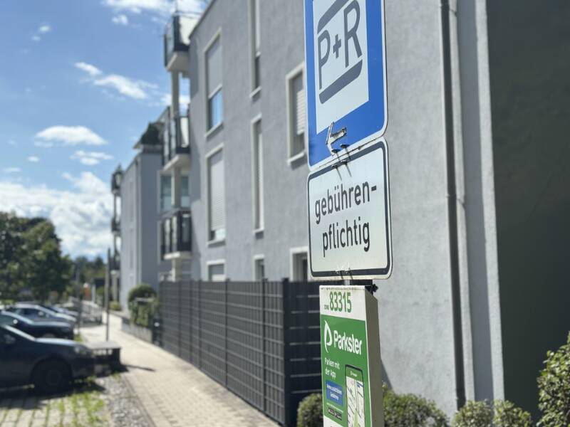 Ein blau-weißes Schild zeigt "P+R" und darunter steht "gebührenpflichtig" in einer städtischen Umgebung mit geparkten Autos und einem Wohngebäude im Hintergrund.