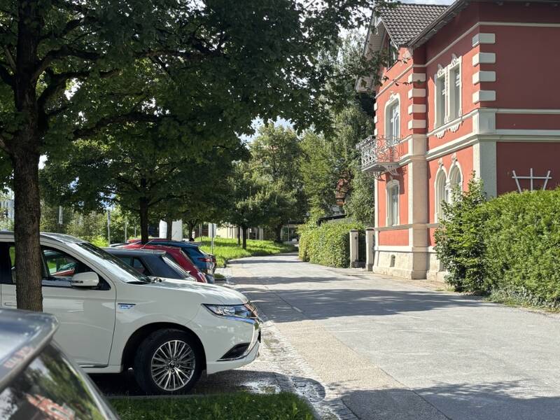 Eine ruhige Straße mit geparkten Autos und einem großen, roten Gebäude neben grünen Bäumen unter blauem Himmel.