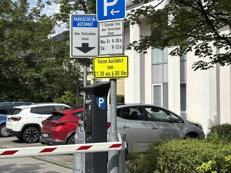 Ein Parkplatz mit mehreren Schildern für Parkscheinautomaten und Ausfahrtszeiten, umgeben von geparkten Autos, einer Schranke und Bäumen.
