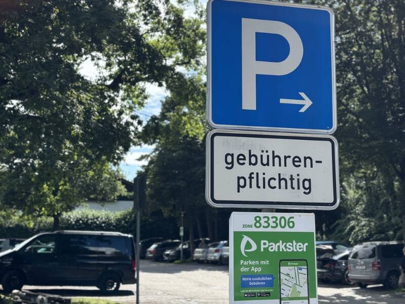 Ein blaues Parkschild weist auf gebührenpflichtiges Parken mit einer App hin, umgeben von Bäumen und parkenden Autos im Hintergrund.