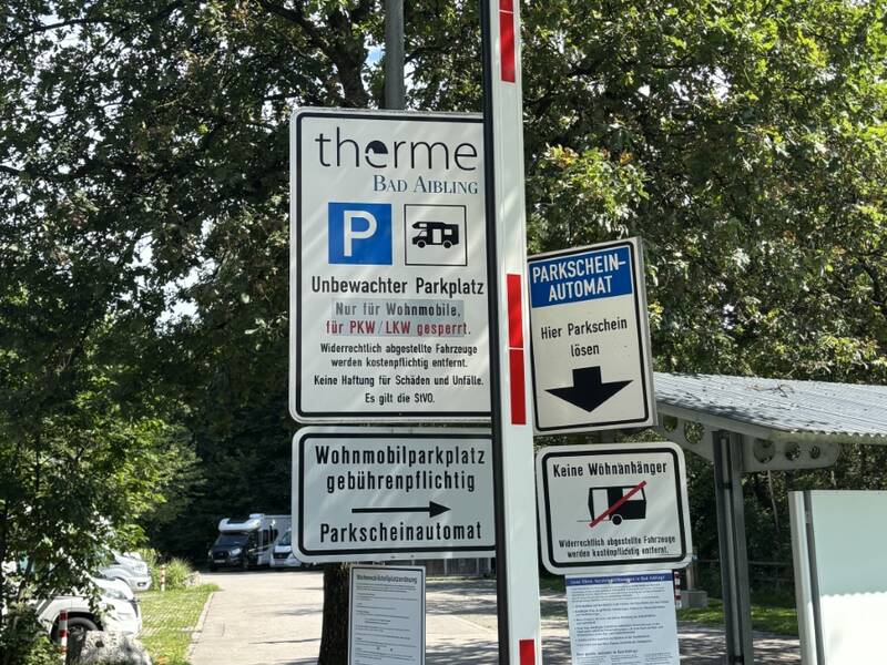 Vor einer grünen Waldkulisse befindet sich ein Schild mit Informationen über einen unbewachten Parkplatz für Wohnmobile in der Nähe der Therme Bad Aibling, ein Parkscheinautomat und Hinweise, dass keine Wohnanhänger erlaubt sind.