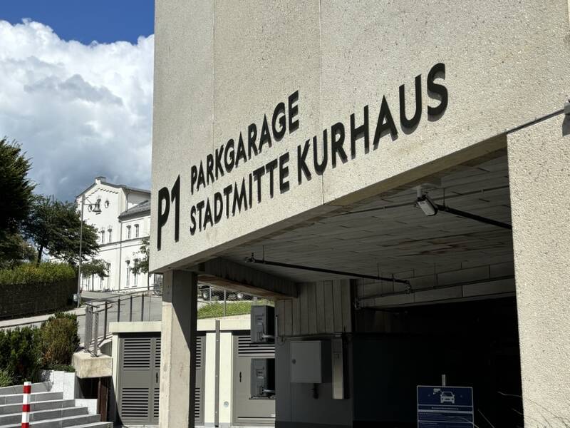 Das Bild zeigt den Eingang zur "Parkgarage Stadtmitte Kurhaus" mit einem Hinweisschild darüber, umgeben von blauem Himmel und grüner Vegetation.